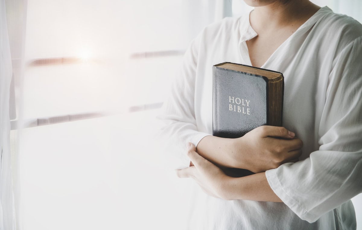 standing-firm-gods-help Image of woman standing by window, holding her Bible in crossed arms.